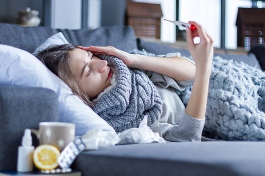 A woman with flu lies on the couch and looks at a fever thermometer.