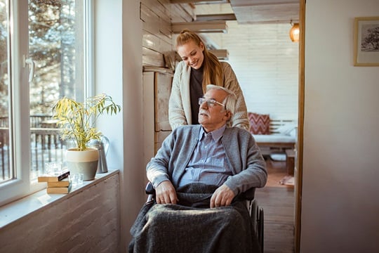 A young woman pushes an elderly man in a wheelchair in his home.