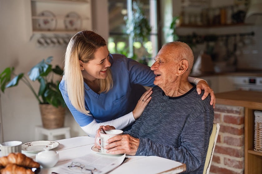 A young woman hugs an older man sitting at the dining table. They are both smiling.