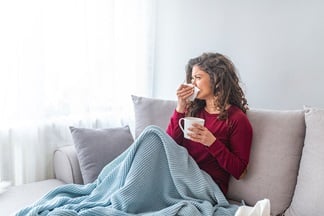 Woman with flu sits on the couch wrapped in a blanket and blows her nose.