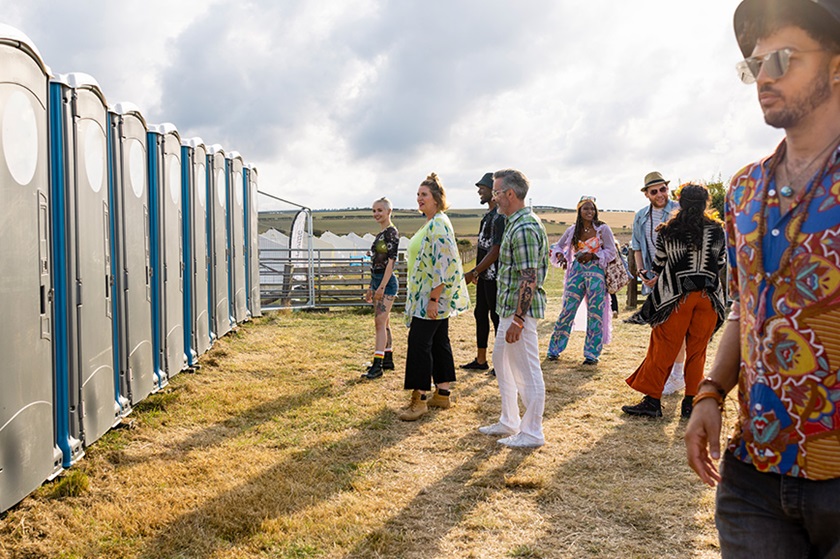 Several people queue up in front of porta-potties at a festival.