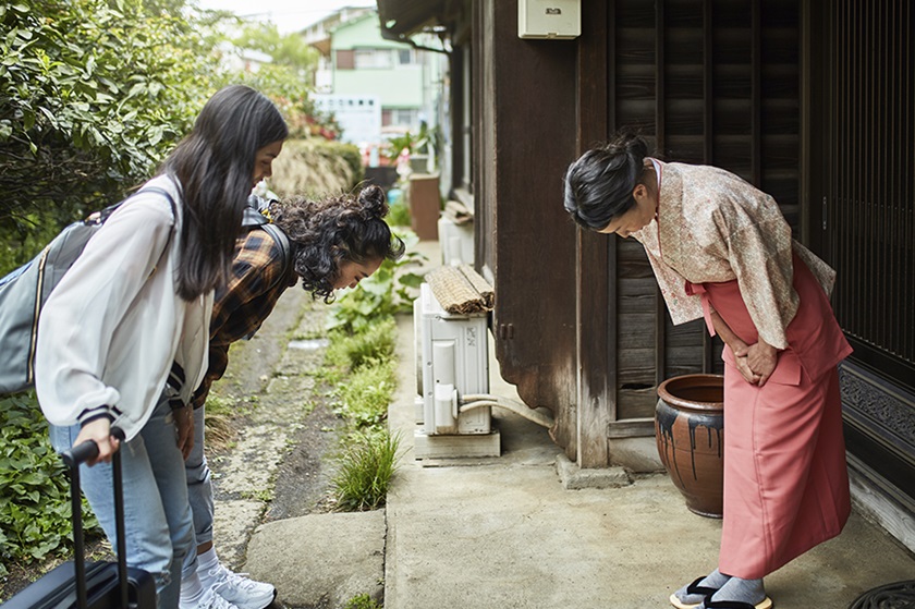 Two young women bow in greeting to an older woman in traditional Japanese garb.