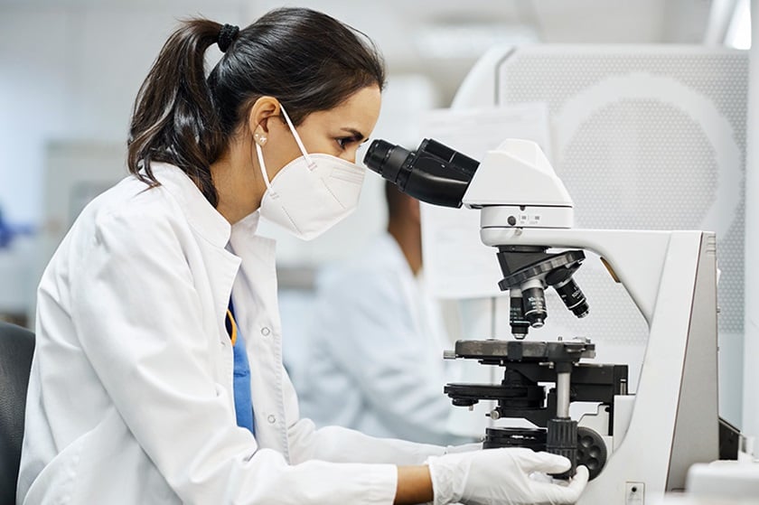 A researcher in a white coat looks into a microscope.